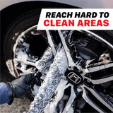 A close-up of a car wheel being washed with foam, showing a gloved hand using a microfiber wheel wash pad on the wheel spokes. A white text banner at the top reads 'REACH HARD TO CLEAN AREAS' in black and red. 112000WS Microfiber Wheel Scrubbing Belt - 2 Pack
