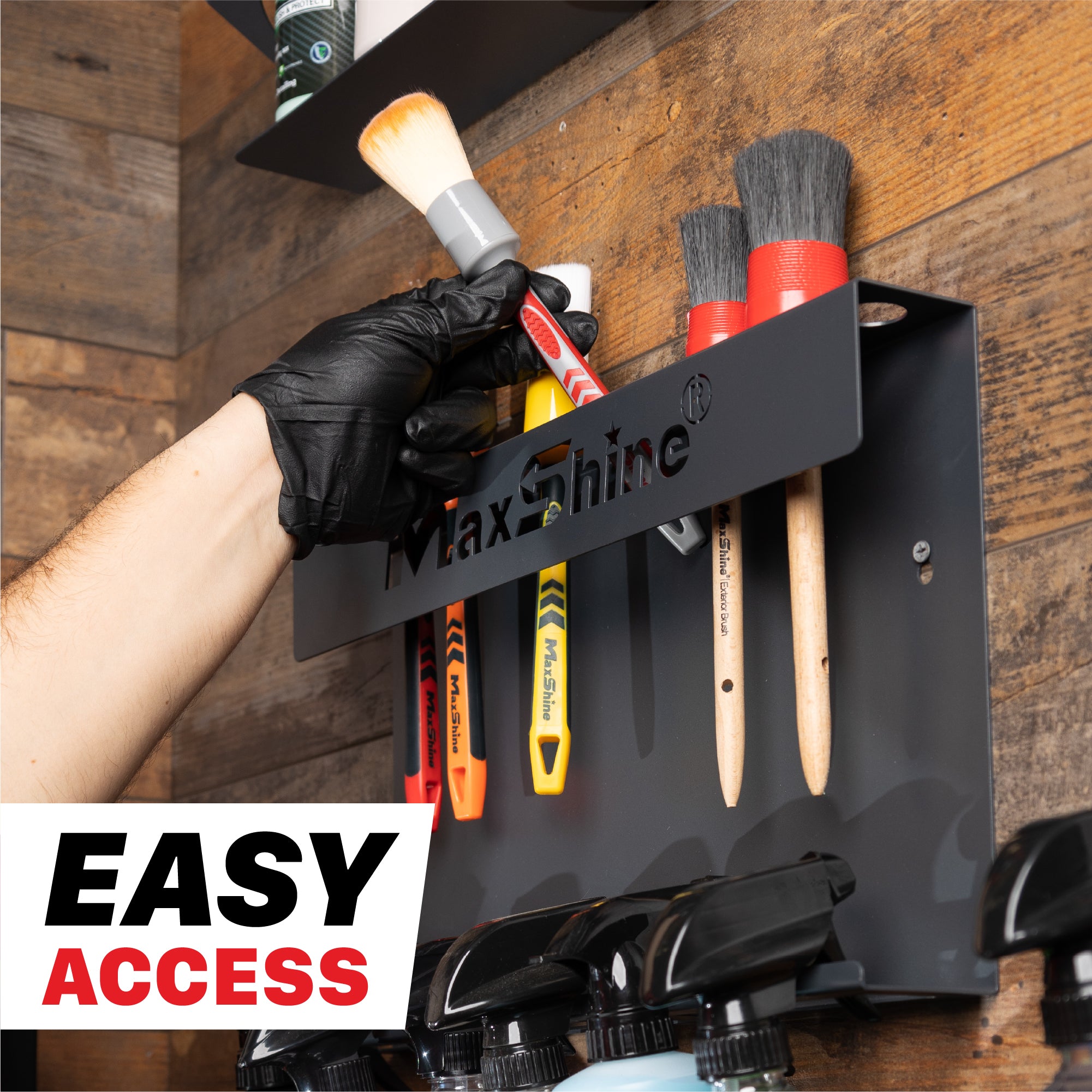A black MaxShine wall-mounted brush holder with several detailing brushes and spray bottles hanging on it, shown in a real environment against a wood-paneled wall. A gloved hand is reaching for one of the brushes, and a white badge reading 'EASY ACCESS' appears in the lower left corner. H06 Wall Mount Tool Holder